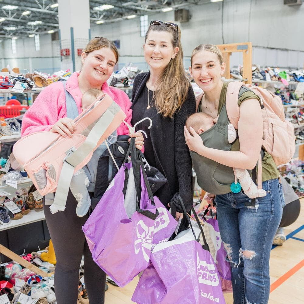 Three friends shop together at their local JBF sale. Mom carries baby in a baby carrier.