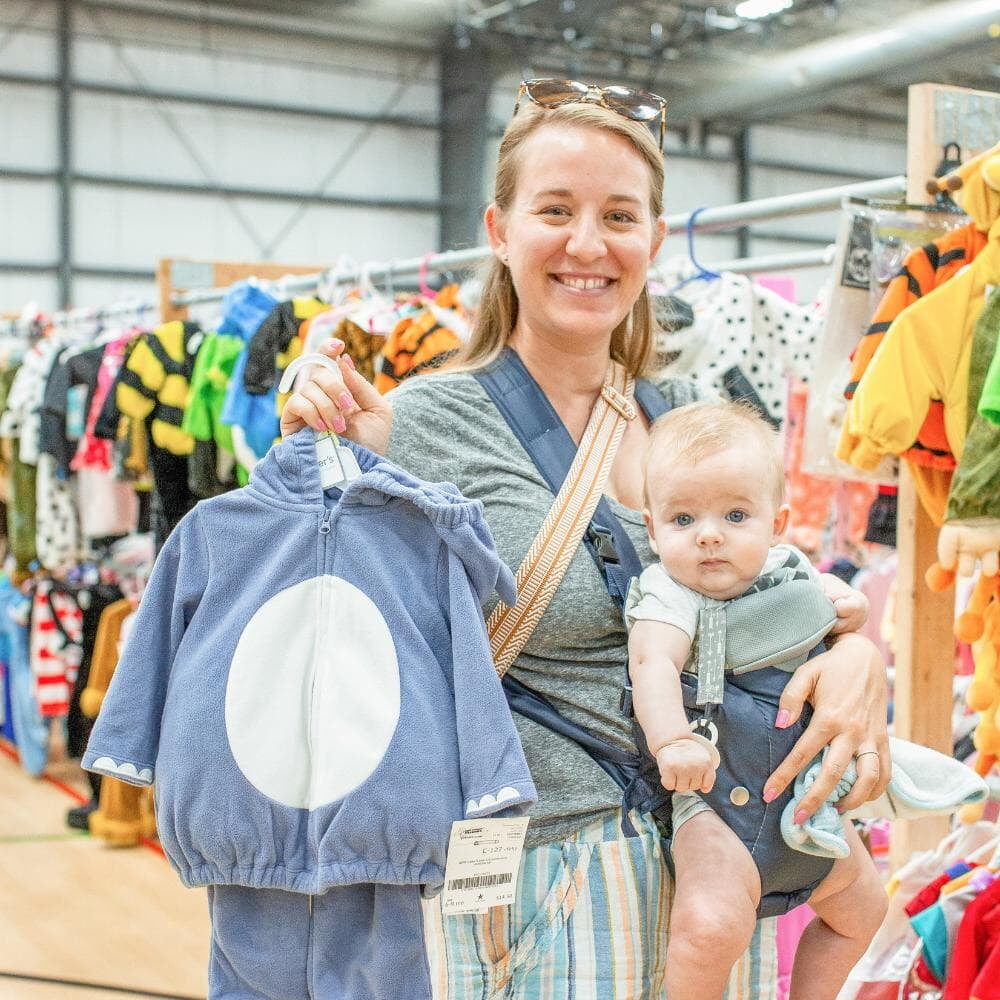 A mom and baby stand beside a rack of clothing at a JBF sale in Texas.